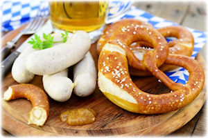 Typische Brotzeit in Bayern: Bier, Weißwurst, Senf und Bretzel. Ein Holzbrett, auf dem verschiedene Nahrungsmittel liegen. Daneben ein Bier.